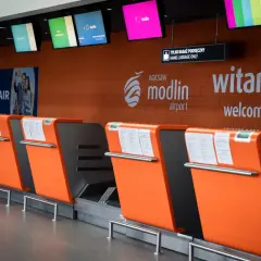 Pomarańczowe stanowiska odprawy bagażowej na lotnisku Modlin z tablicami informacyjnymi i logo lotniska.