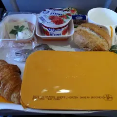 Taca z jedzeniem w samolocie: rogalik, jogurt, bułka, masło, kawa i żółty pojemnik.