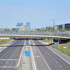 Droga ekspresowa z wiaduktem i widokiem na nowoczesne budynki w tle.