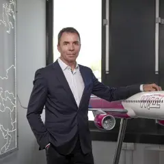 Mężczyzna w garniturze stoi obok modelu samolotu Wizz Air, z mapą Europy w tle.
