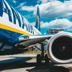 Fragment samolotu Ryanair z widocznym silnikiem i podwoziem na tle błękitnego nieba z chmurami.
