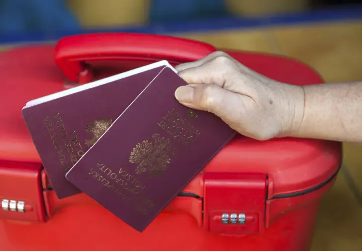Dłoń trzyma polskie paszporty na czerwonej walizce. Kto może odebrać paszport osoby dorosłej? Tylko ona!