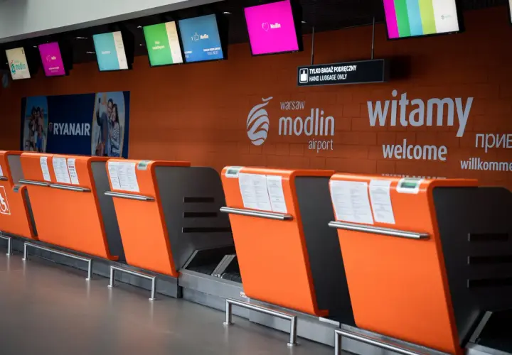 Pomarańczowe stanowiska odprawy bagażowej na lotnisku Modlin z tablicami informacyjnymi i logo lotniska.