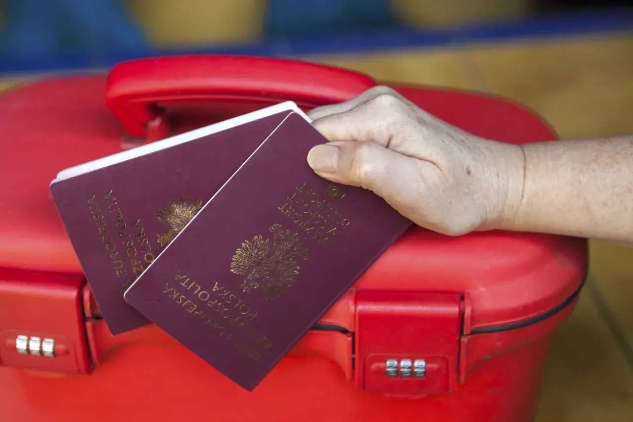 Dłoń trzyma polskie paszporty na czerwonej walizce. Kto może odebrać paszport osoby dorosłej? Tylko ona!
