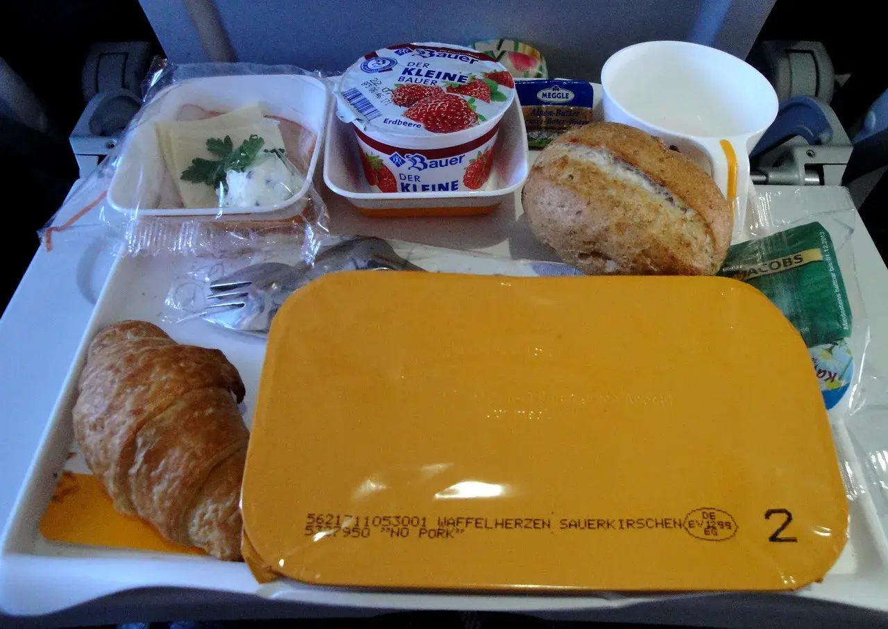 Taca z jedzeniem w samolocie: rogalik, jogurt, bułka, masło, kawa i żółty pojemnik.