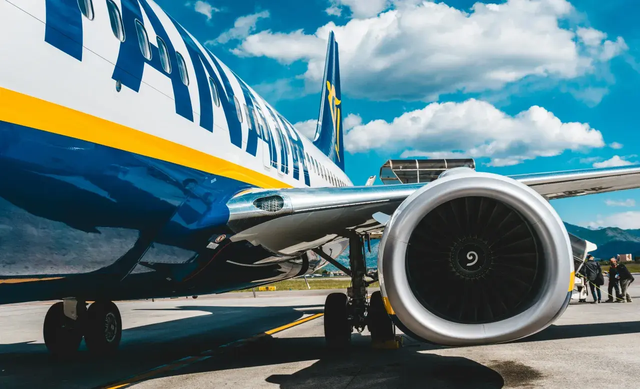 Fragment samolotu Ryanair z widocznym silnikiem i podwoziem na tle błękitnego nieba z chmurami.