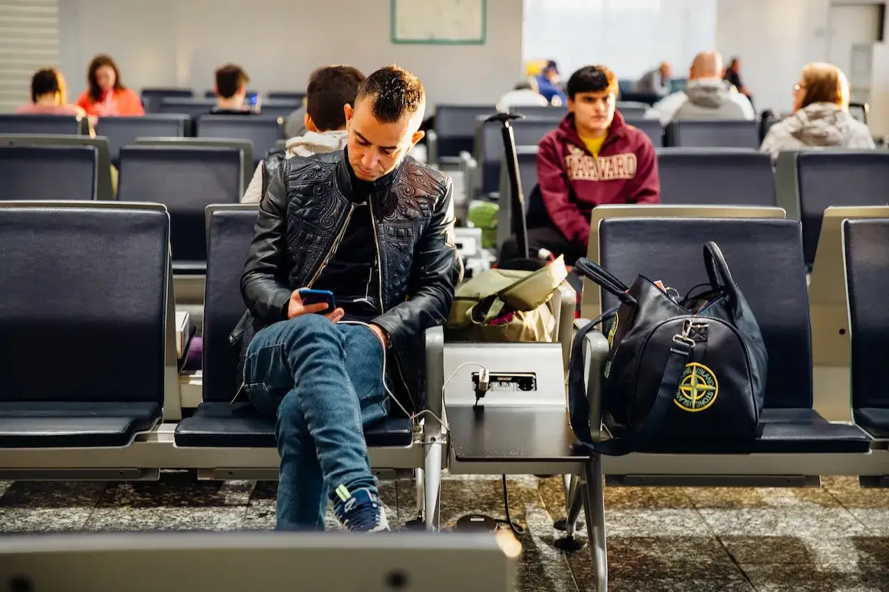 Mężczyzna w czarnej kurtce siedzi na lotnisku, przeglądając telefon. Obok niego torba, może zastanawia się, jak dokupić bagaż do lotu.